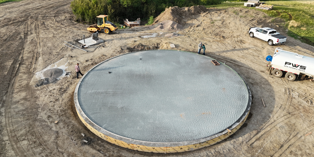 Aerial view of a circular concrete tank under construction, with two workers standing on the slab, construction equipment nearby, and a white truck labeled “PWS” parked at the edge of the site.