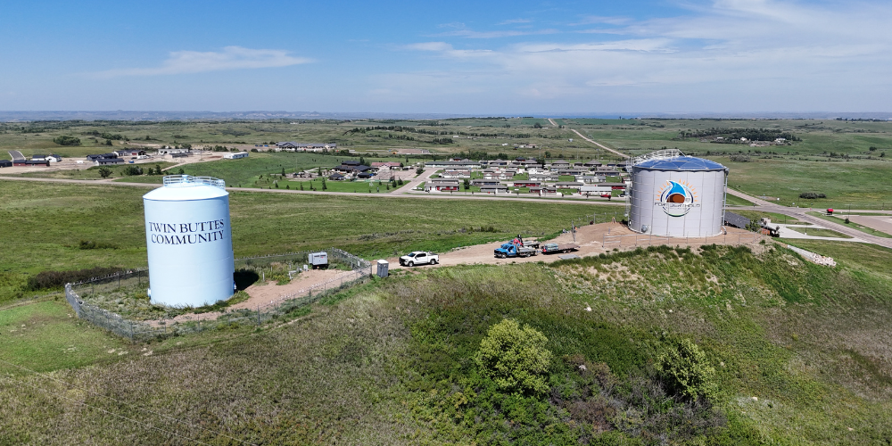 Aerial view of two water towers labeled “TWIN BUTTES COMMUNITY” overlooking a small town and surrounding prairie.