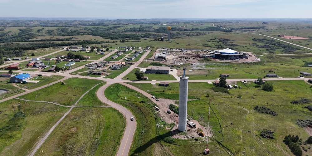 Aerial view of a tall concrete water tank under construction in the foreground, with a road curving past it and a small community, construction site, and rolling green landscape visible in the background.