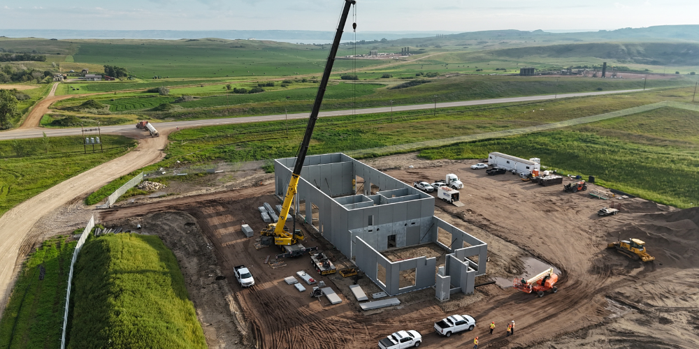 Aerial view of a crane installing components on a concrete building under construction in a rural landscape.