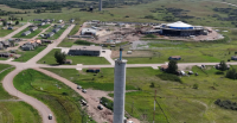 Aerial view of a tall concrete water tank under construction in the foreground, with a road curving past it and a small community, construction site, and rolling green landscape visible in the background.