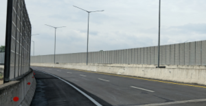 A curved highway lined with tall, gray noise‑barrier walls and streetlights under a partly cloudy sky.