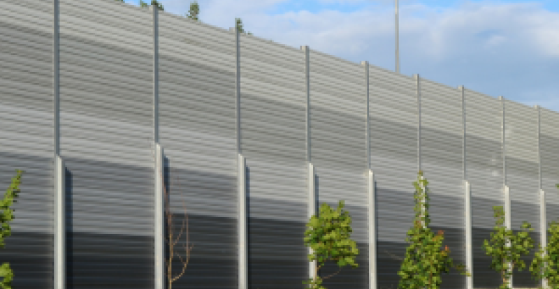 A row of young trees planted along a grassy embankment in front of a tall, gray metal noise‑barrier wall.