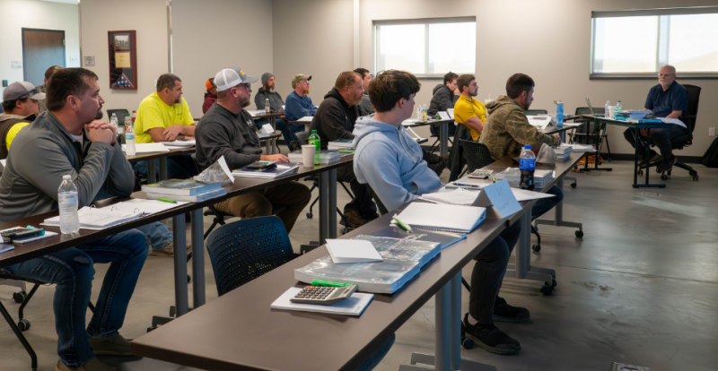 Group of professionals attending a classroom training, seated at tables and listening to an instructor at the front.