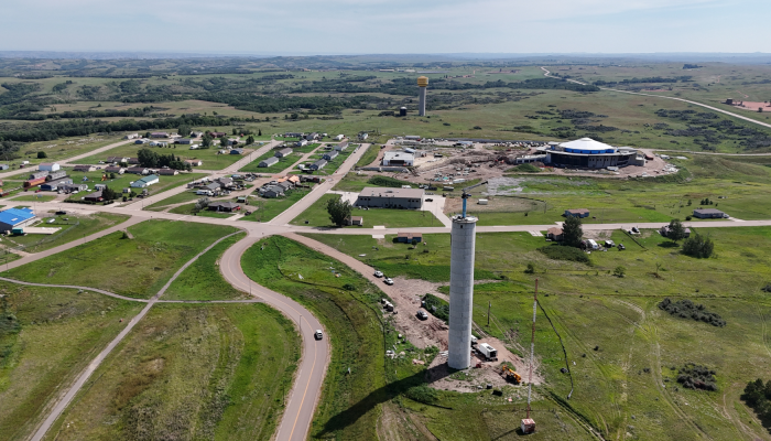 Aerial view of a tall concrete water tank under construction in the foreground, with a road curving past it and a small community, construction site, and rolling green landscape visible in the background.